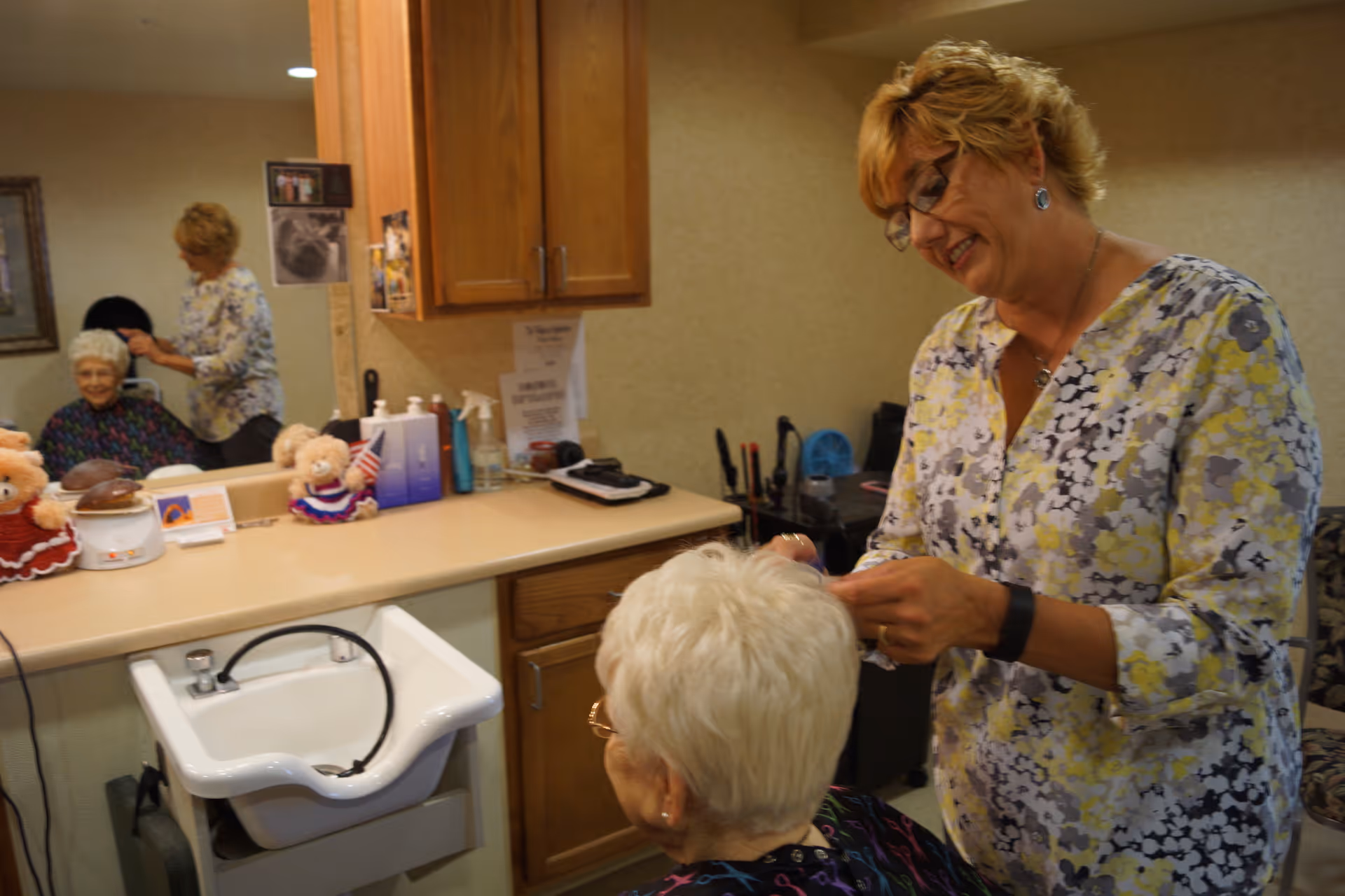 A hairdresser styling the hair of an elderly woman seated in a chair in a salon area. The room has a countertop with various hair care products, a small sink, and a mirror reflecting the scene. The hairdresser is smiling and wearing glasses and a floral patterned blouse.