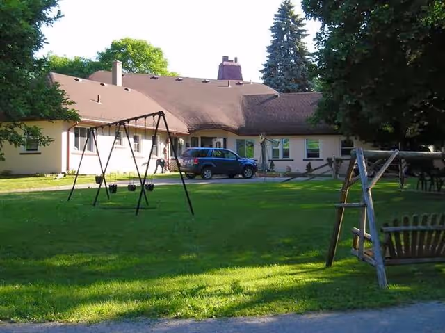 Single-story light-colored building with a green lawn, playground swings, a parked SUV, and trees in the front yard.