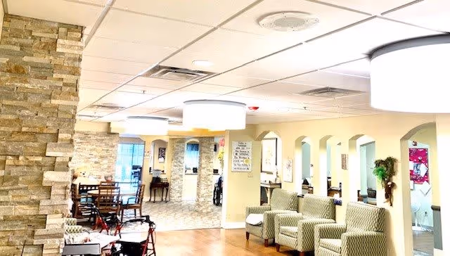A bright and spacious interior common area in a senior living facility with patterned armchairs lined up against a wall, a stone accent wall, wooden chairs and tables, and large ceiling lights. The space has a warm and welcoming atmosphere with light-colored walls and flooring.