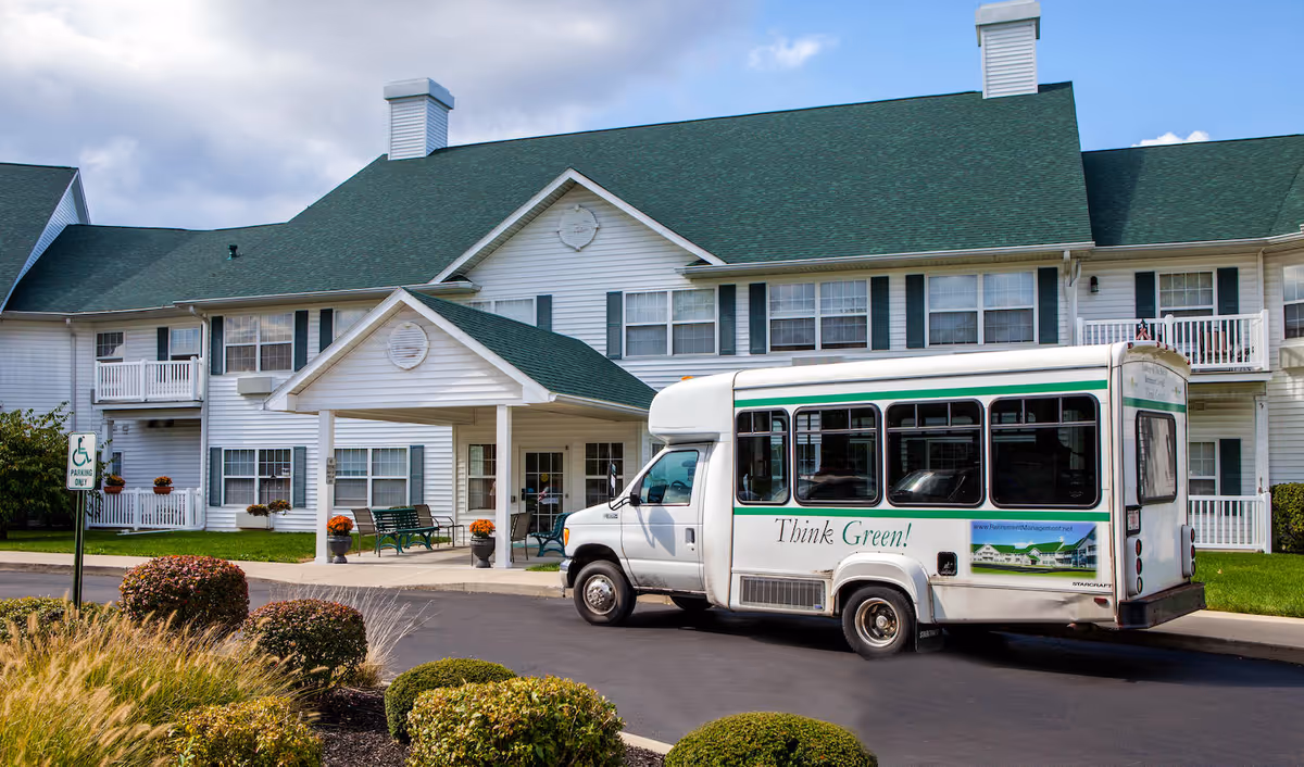 Exterior view of Northwood Manor, a two-story senior living facility with white siding and green roof. A white shuttle bus with the words 'Think Green!' is parked in front of the entrance. There are benches and potted plants near the entrance, and landscaped bushes and grass around the driveway.