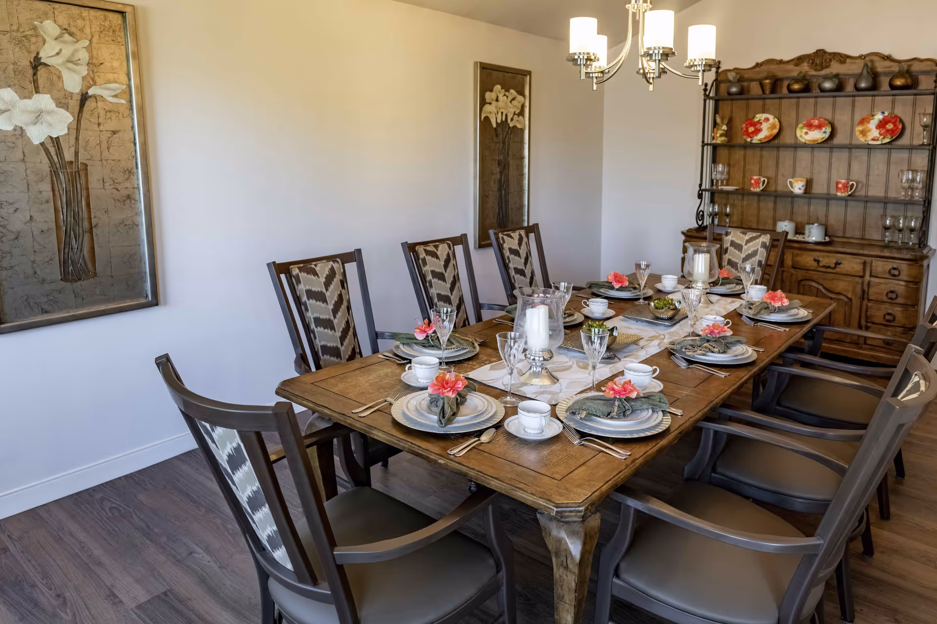 A dining room with a wooden table set for eight people. Each place setting includes plates, cups, silverware, and a napkin with a pink flower. The chairs have patterned backs and cushioned seats. A wooden hutch with decorative plates and glassware is against the wall, and two framed floral artworks hang on the walls. A chandelier with five lights hangs above the table.