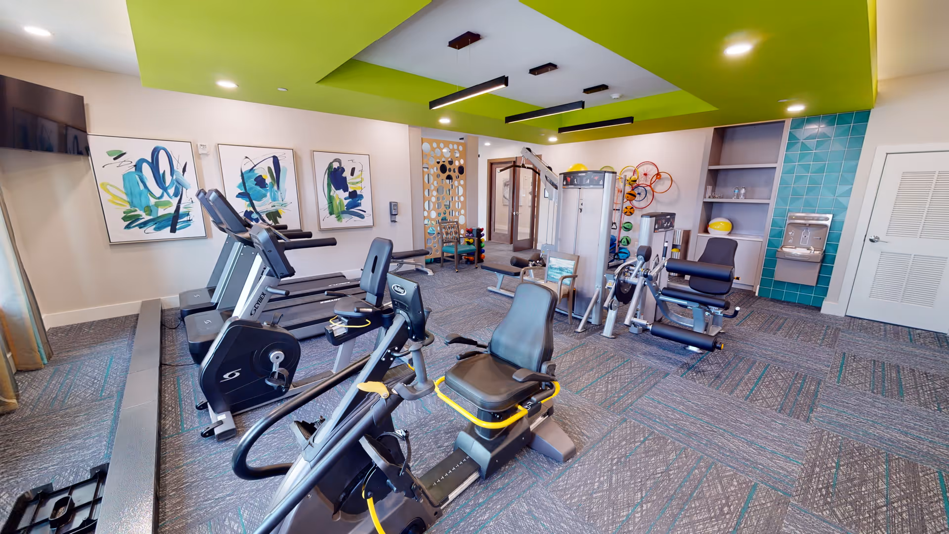 A senior living facility fitness room with exercise equipment including stationary bikes, treadmills, and weight machines. The room has a green ceiling accent, abstract artwork on the walls, a water fountain, and a carpeted floor.