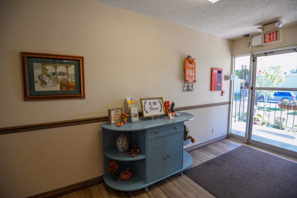 Entrance area of a facility with a light blue console table decorated with small pumpkins, flowers, and framed signs including one that says 'Be Our Guest'. A framed picture hangs on the wall above the table, and a 'Happy Fall Y'all' sign is mounted near the exit door. The door is glass with an exit sign above it, showing a view of the outside parking area and some greenery.