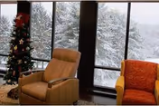 A cozy indoor seating area with two armchairs, one beige and one orange, positioned near large windows showing a snowy outdoor scene with snow-covered trees. A decorated Christmas tree is visible on the left side of the image.