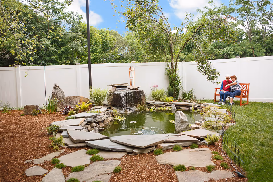 Two elderly women sitting on a wooden bench in a garden area next to a small pond with a waterfall feature, surrounded by rocks, plants, and a white fence with trees in the background under a partly cloudy sky.