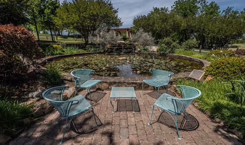Outdoor seating area with five light blue metal chairs arranged around a small matching table on a brick patio. In the background, there is a circular pond with lily pads and a small waterfall feature, surrounded by greenery and trees under a partly cloudy sky.
