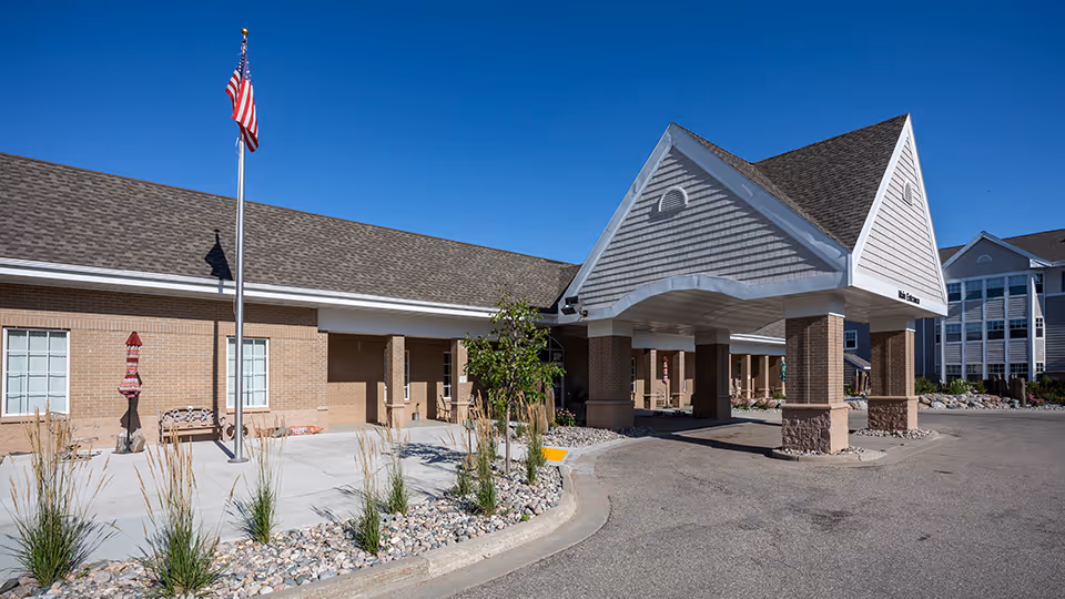 Exterior view of Valley Senior Living on 42nd - Woodside Village showing the main entrance with a covered drop-off area supported by brick columns, a flagpole with an American flag, landscaping with rocks and plants, and a clear blue sky.