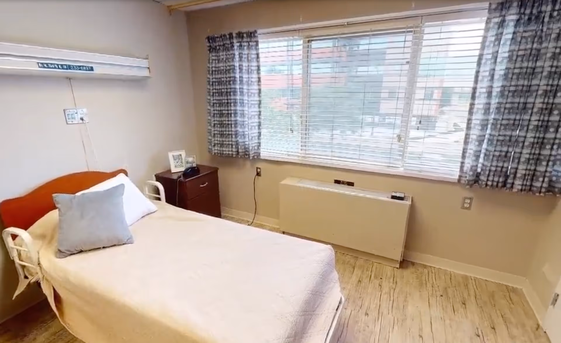 A small, simple bedroom with a single bed covered in a beige blanket and two pillows. Next to the bed is a wooden nightstand with a framed photo and a telephone. A large window with blinds and patterned curtains lets in natural light. The room has light-colored walls and wood-patterned flooring.