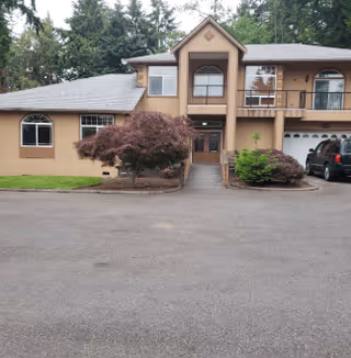 Front exterior view of a two-story residential building with a beige facade, multiple windows, a balcony, and a driveway with parked cars. There are landscaped bushes and trees around the entrance.