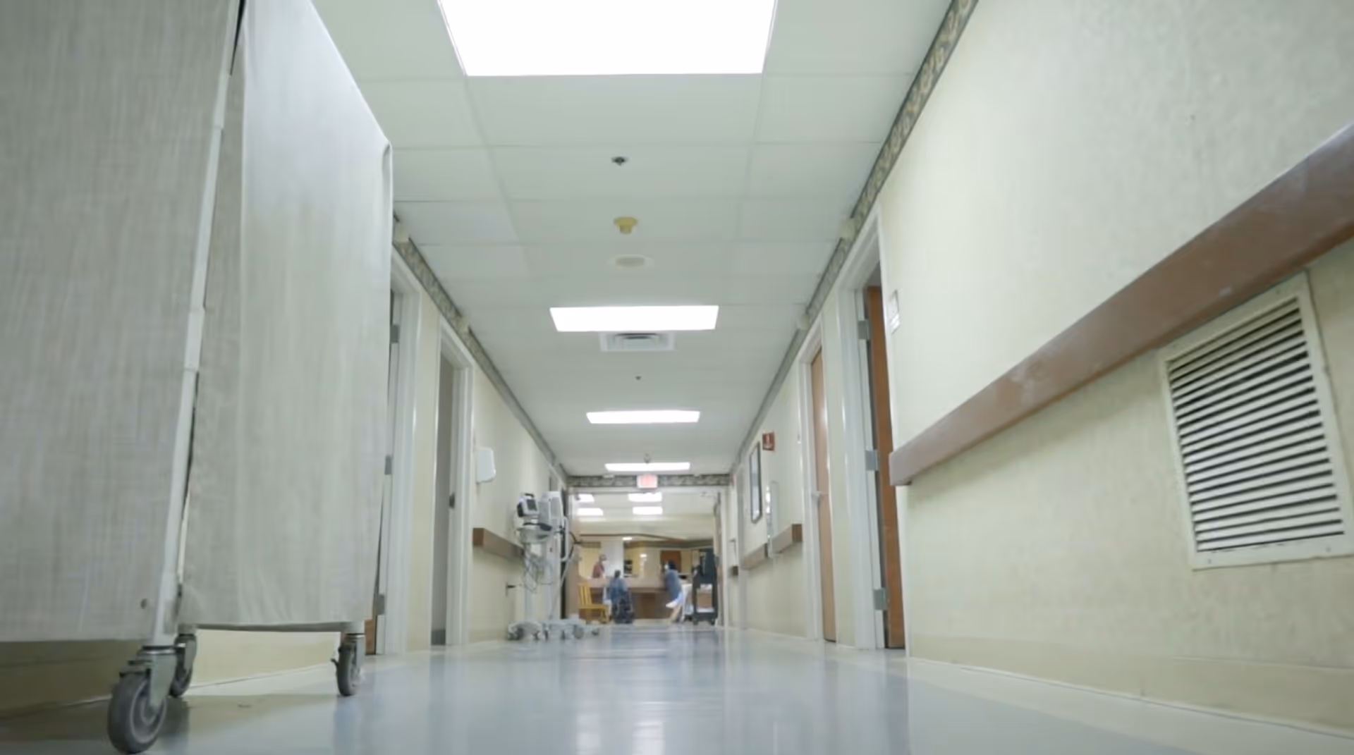Low-angle view of a long, clean hallway in a rehabilitation and nursing center with beige walls, ceiling lights, handrails, and a privacy screen on wheels on the left side. Two people are visible in the distance near the end of the hallway.