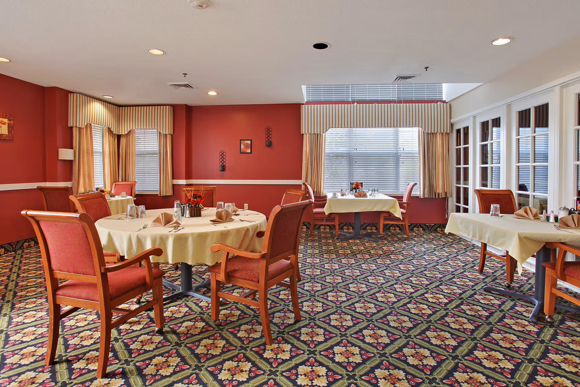 A dining room with red walls and patterned carpet featuring floral designs. There are several tables covered with beige tablecloths, each set with napkins, glasses, and silverware. The room has large windows with beige curtains and valances, and wooden chairs with red upholstery surround the tables.