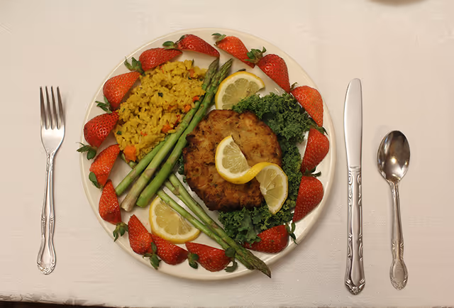 Plated meal of a crab cake with lemon slices, asparagus, yellow rice, kale and strawberries arranged on a white plate with fork, knife and spoon on a white tablecloth.