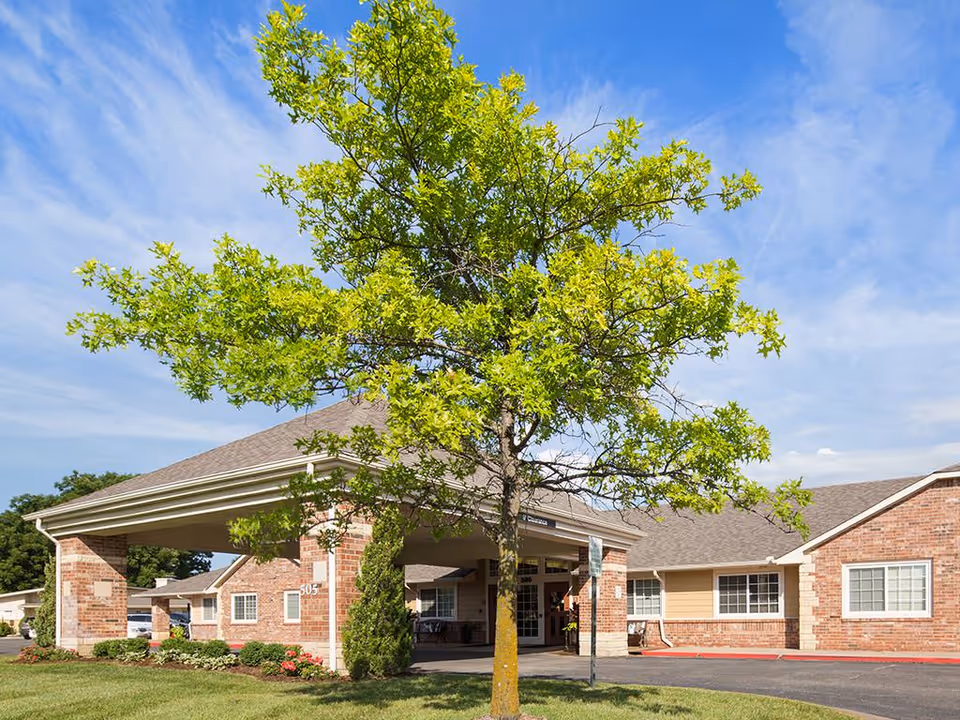 Front entrance of a single-story brick senior living building with a porte-cochere and a leafy tree in the foreground.