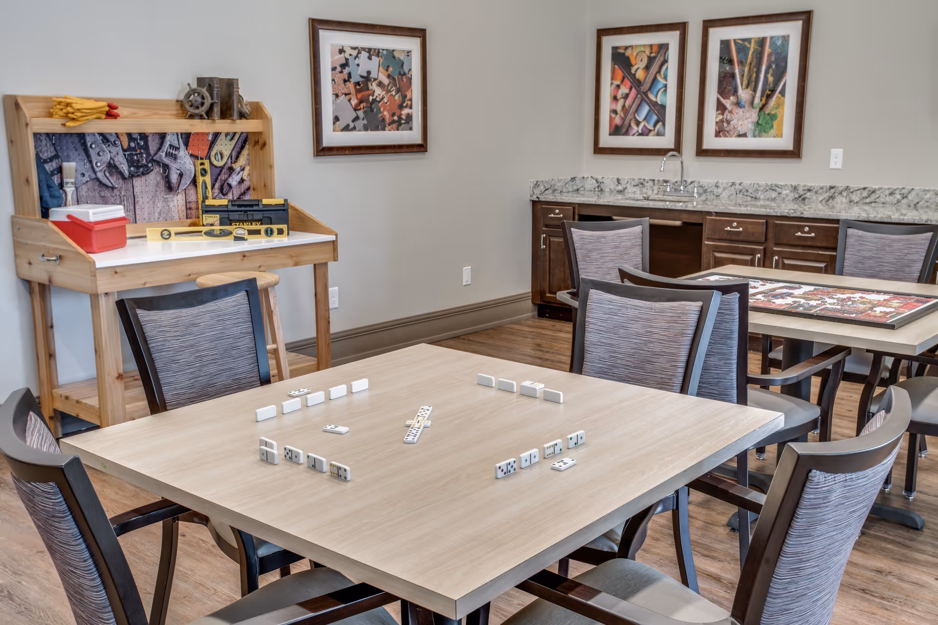 A well-lit activity room with tables and chairs arranged for playing games. One table has dominoes set up for a game, and another table has a partially completed jigsaw puzzle. The room features a wooden workbench with tools and a red toolbox, a countertop with a sink, and three framed pictures on the wall depicting puzzle pieces and art supplies.