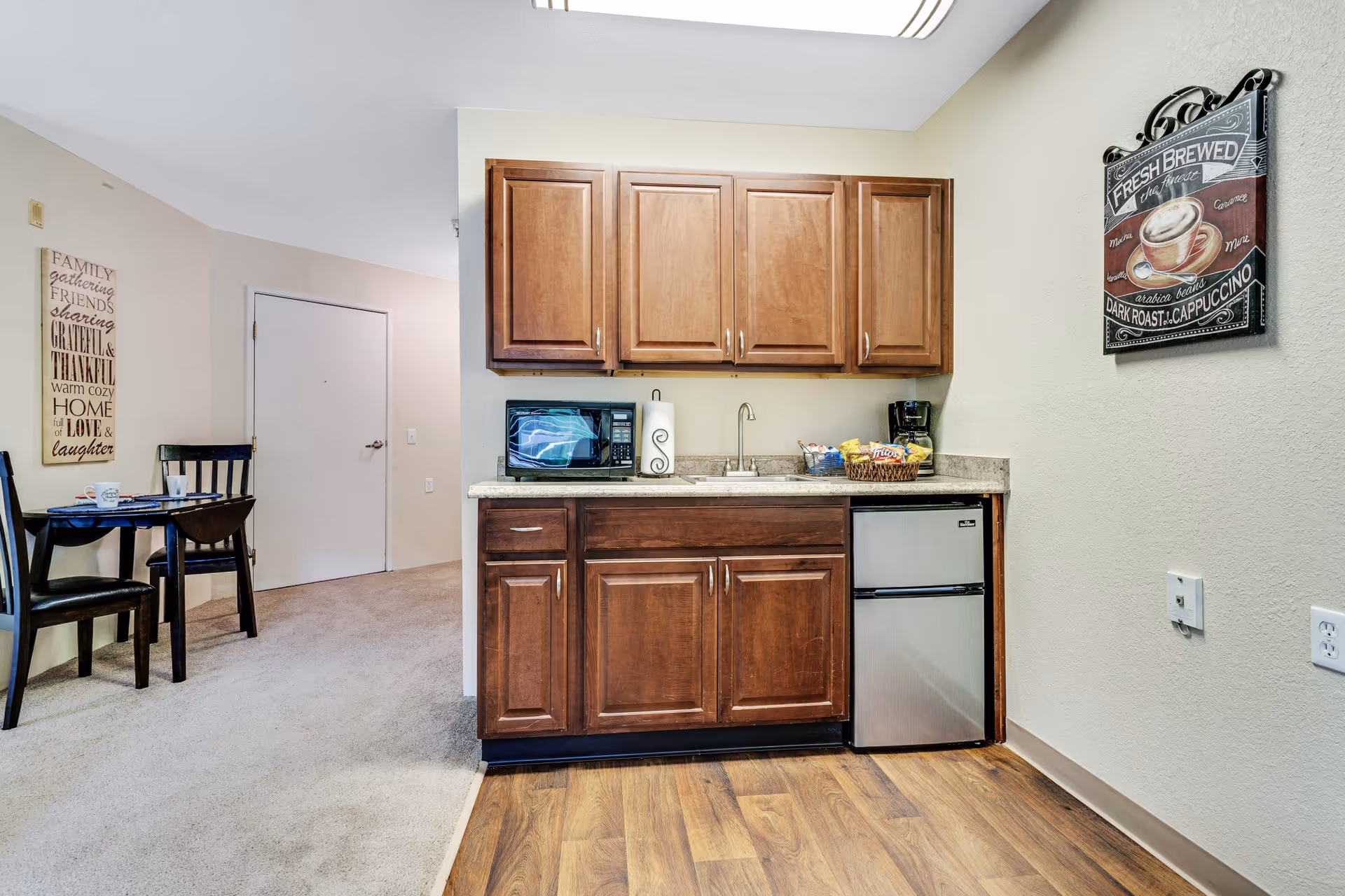 A small kitchenette area with wooden cabinets, a countertop with a microwave, paper towel holder, coffee maker, and baskets of snacks. To the left, there is a small dining table with two chairs and a wall decoration with inspirational words. The floor transitions from wood in the kitchenette to carpet in the dining area.