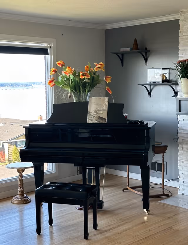 Black grand piano with bench and a vase of orange tulips in a bright living room by a large window.