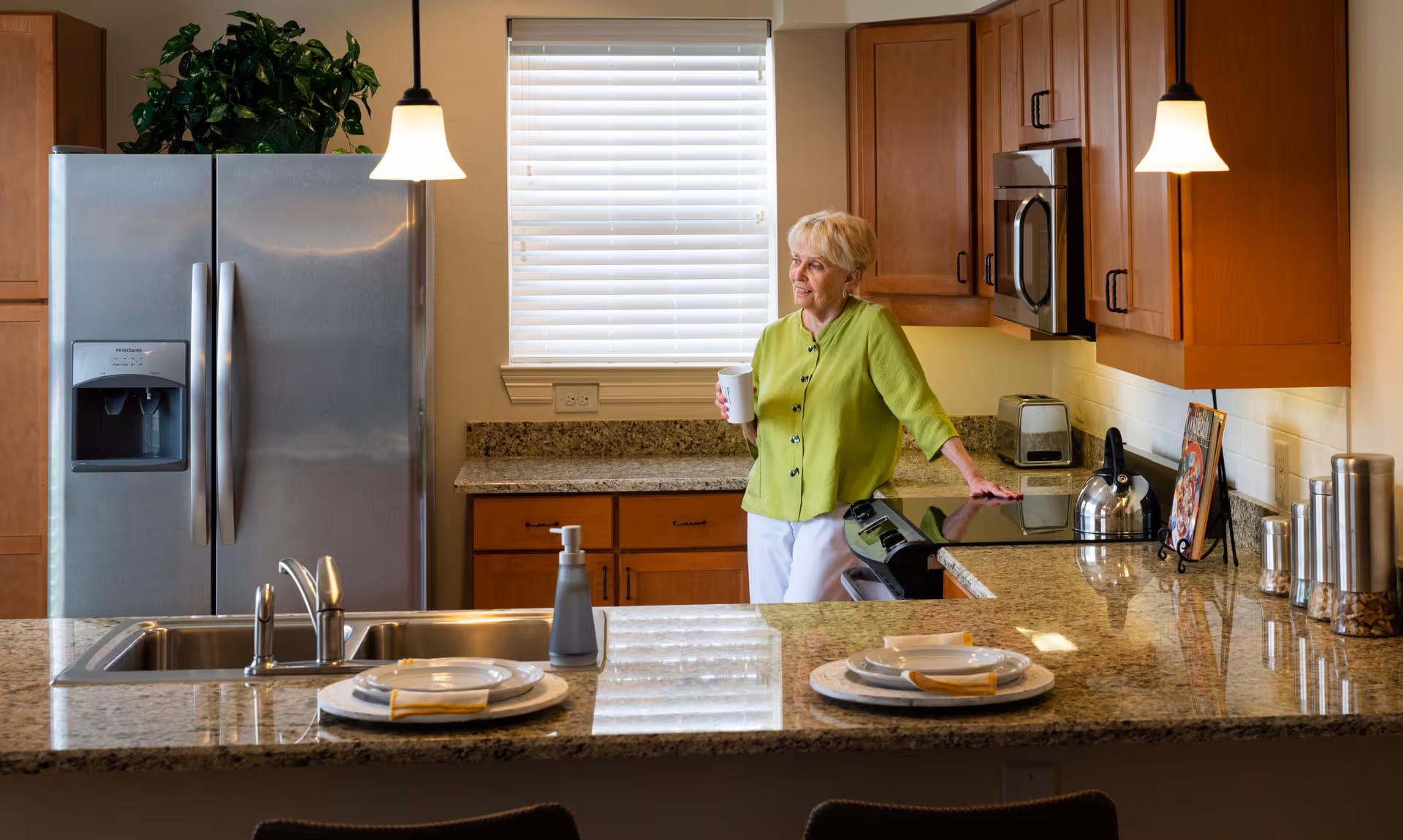 A senior woman in a green blouse and white pants stands in a modern kitchen holding a white cup. The kitchen features granite countertops, wooden cabinets, a stainless steel refrigerator, a sink with a faucet, a stove with a kettle, a toaster, and two pendant lights hanging from the ceiling. There are two place settings on the counter with plates and napkins.