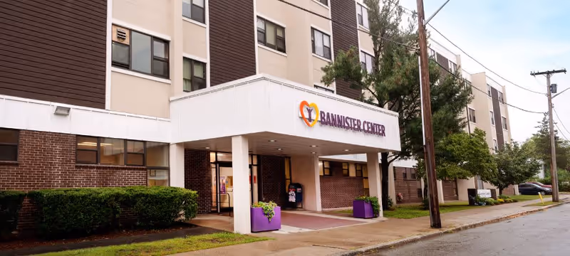 Exterior front entrance of Bannister Center For Rehabilitation And Health Care, a multi-story building with a covered entryway, purple planters with flowers, and surrounding greenery on a cloudy day.