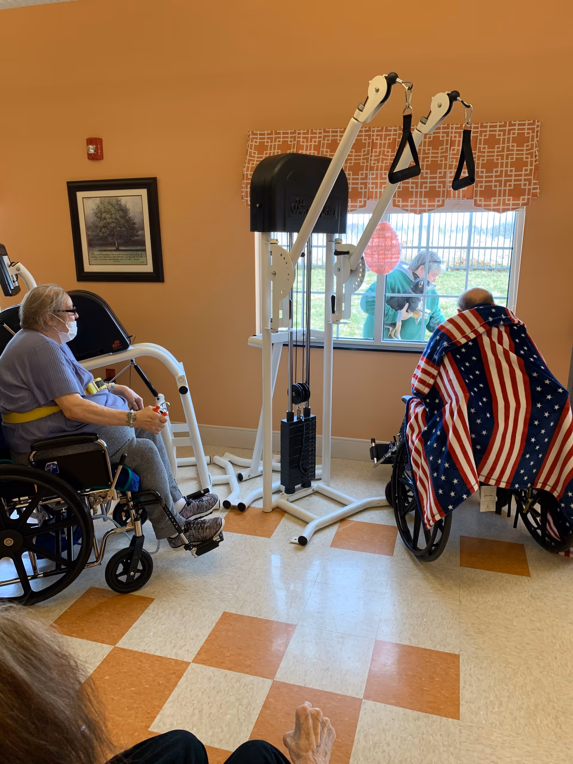 Two elderly individuals in wheelchairs are inside a room with exercise equipment. One person is wearing a mask and exercising with a resistance machine, while the other is covered with an American flag blanket and facing a window. Outside the window, two people are visible, one holding a small dog. The room has a checkered floor and a framed picture on the wall.