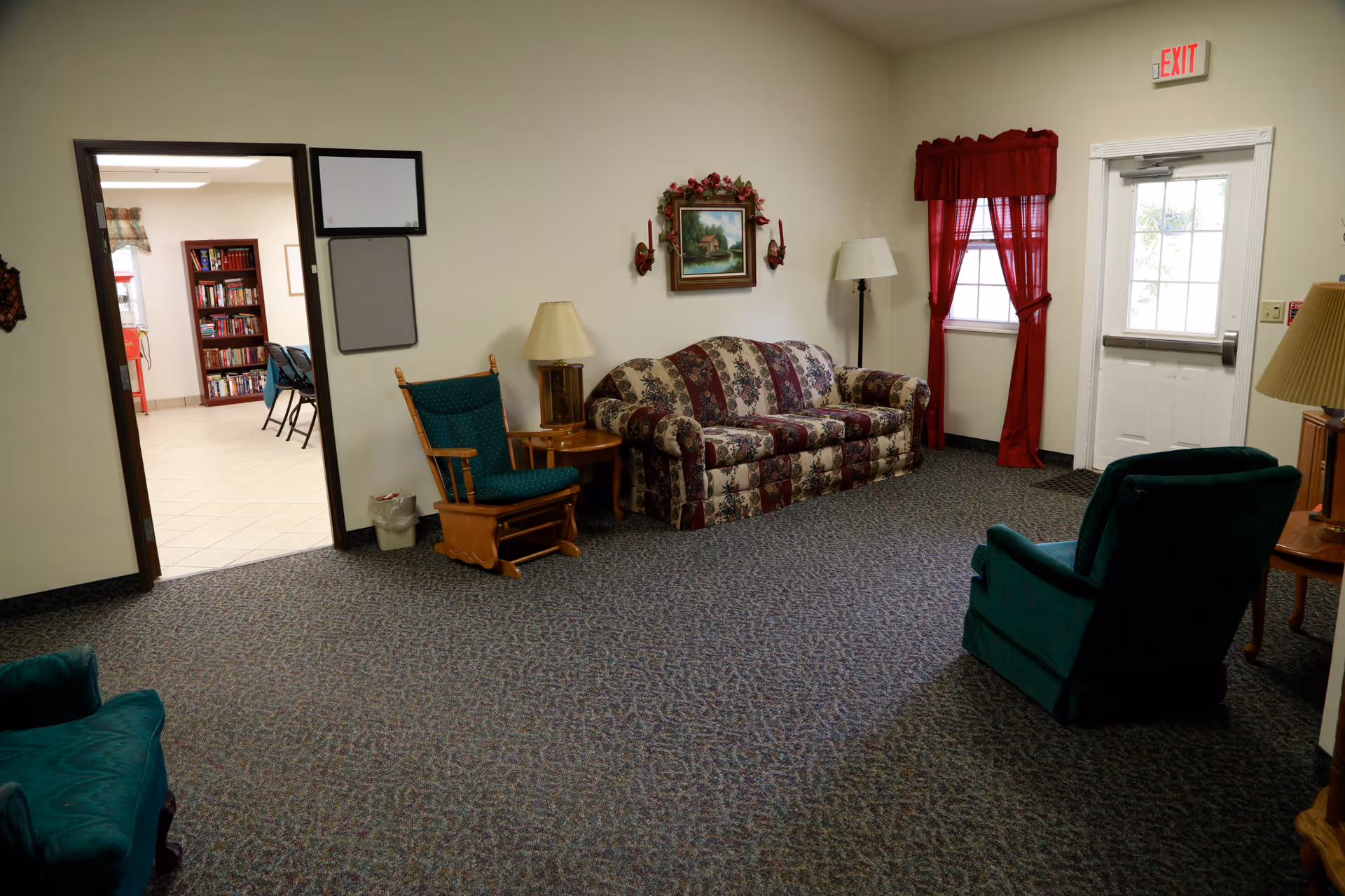A cozy living room area in an assisted living facility featuring a floral patterned sofa, two green upholstered chairs, a wooden rocking chair, two side tables with lamps, a painting on the wall, and a window with red curtains next to a white exit door. An open doorway leads to another room with bookshelves and chairs.