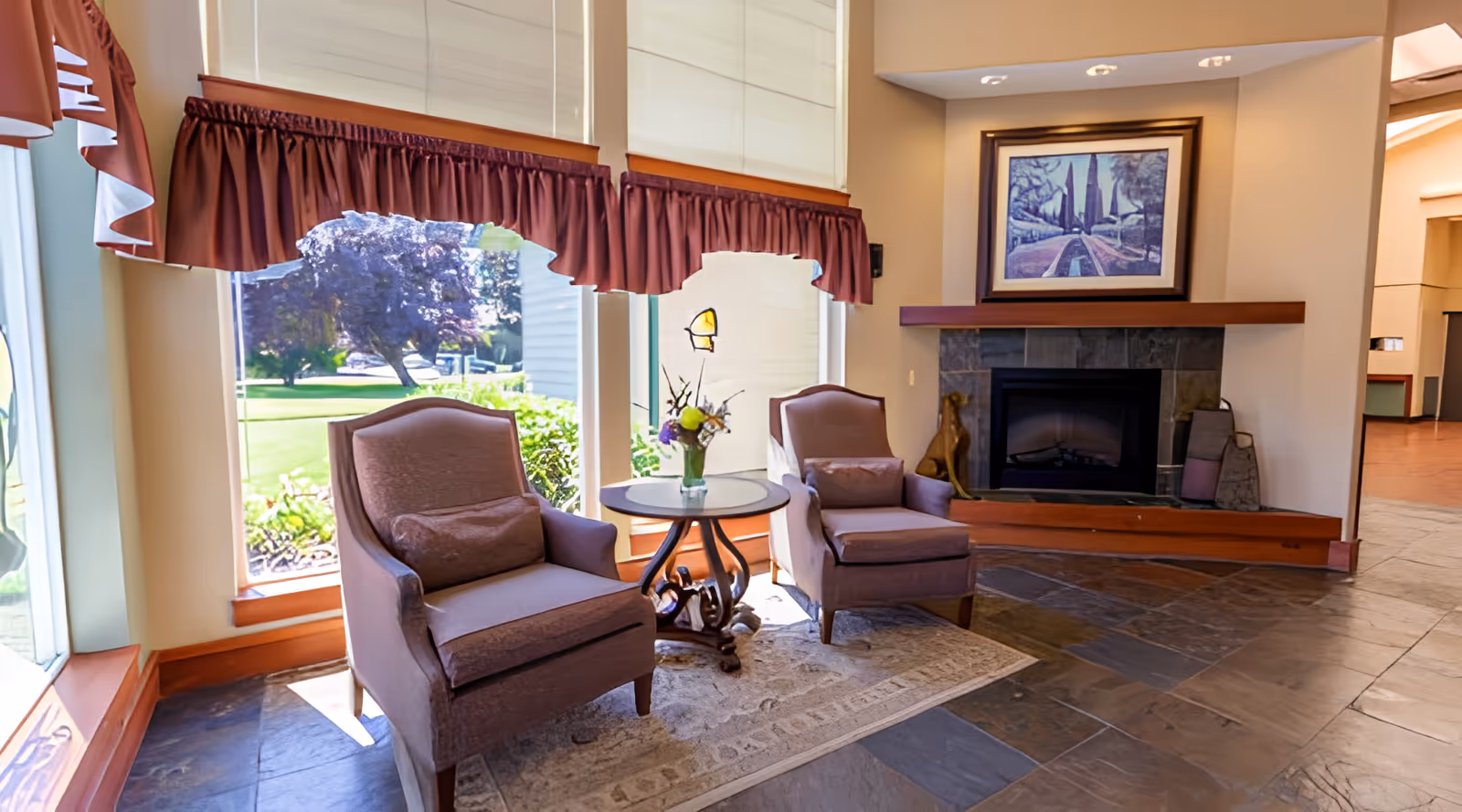 A cozy seating area with two upholstered armchairs and a small round table with a flower vase between them, positioned in front of large windows with maroon valances. To the right is a stone fireplace with a framed artwork above it. The floor is tiled with a stone pattern, and a light-colored rug is placed under the chairs and table. Outside the windows, greenery and trees are visible.
