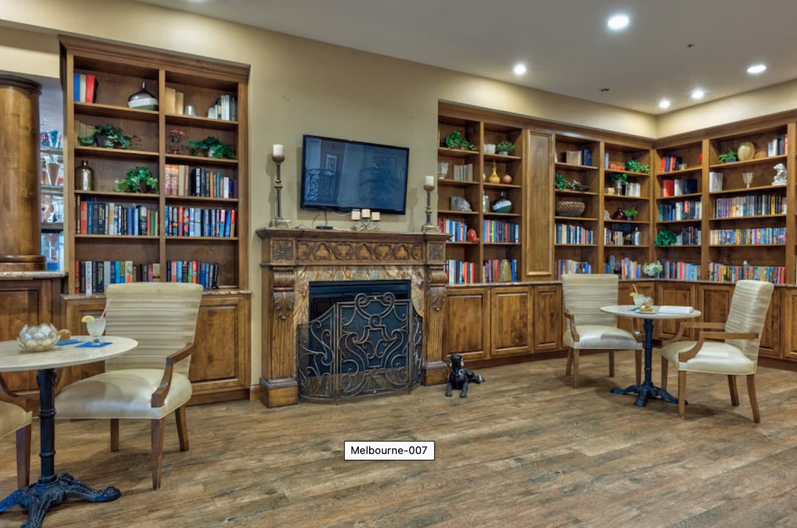 A cozy library or reading room with wooden bookshelves filled with books and decorative plants. There is a fireplace with an ornate wooden mantle and a metal screen in front. Above the fireplace is a flat-screen TV. Two small round tables with cream-colored upholstered chairs are placed in the room, each table holding drinks and small decorative items. The floor is wooden, and the lighting is warm and inviting.