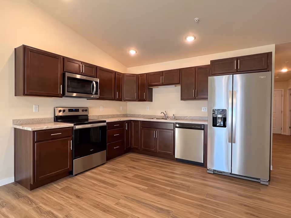 Modern kitchen with dark wood cabinets, stainless steel appliances including a microwave, oven, dishwasher, and refrigerator, and wood flooring under warm ceiling lights.