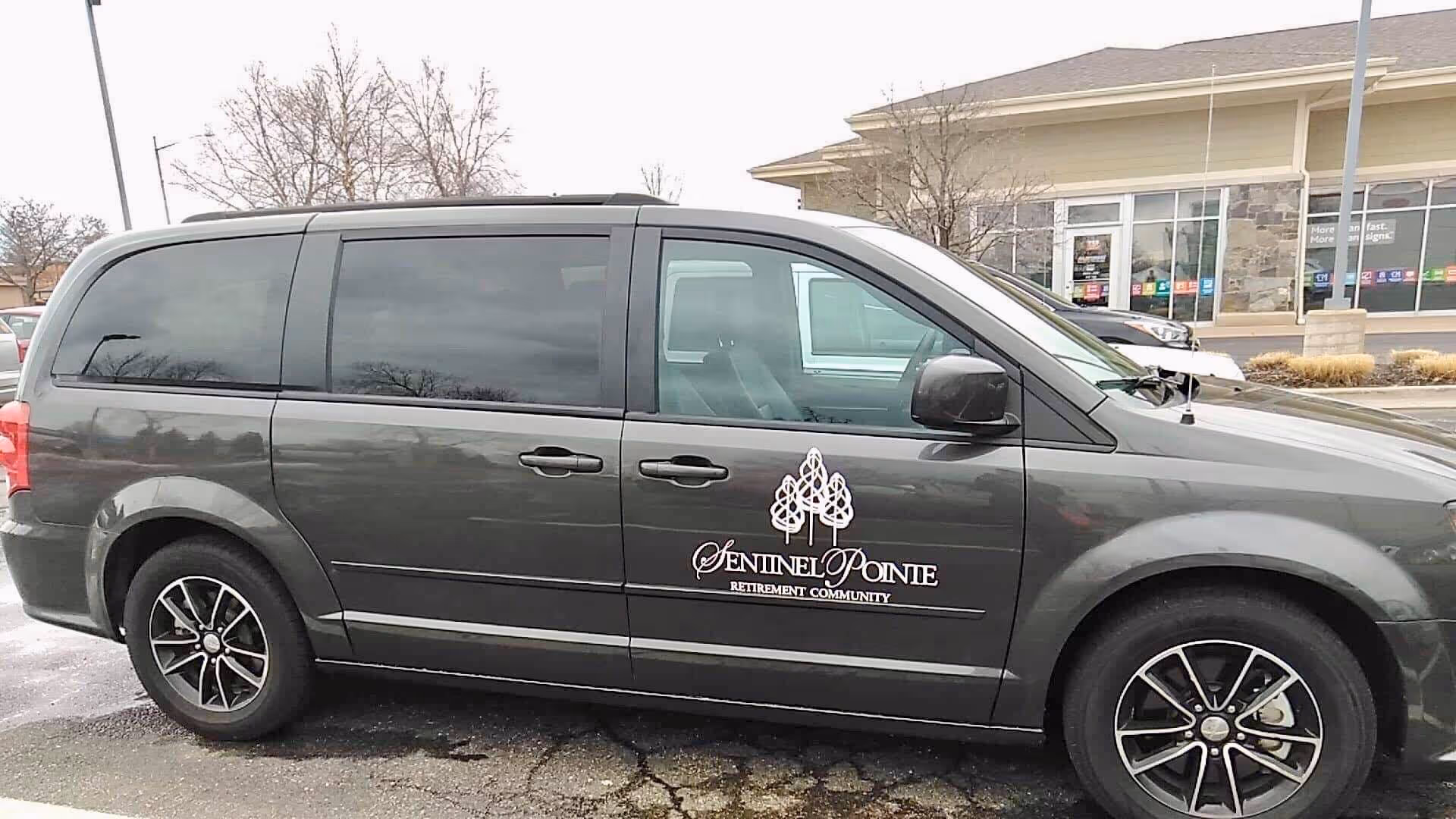 A gray minivan parked in a parking lot with the logo and text 'Sentinel Pointe Retirement Community' on the side door. The background shows a commercial building with large windows and some leafless trees.