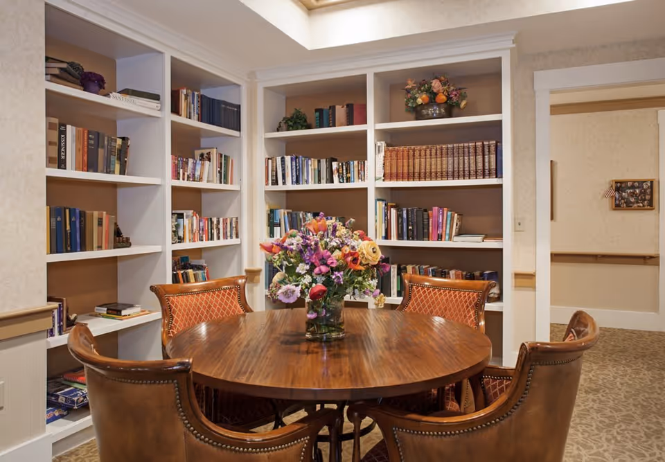 A cozy library room with white built-in bookshelves filled with books and decorative items. In the center, there is a round wooden table with a vase of colorful flowers and four upholstered chairs around it. The room has warm lighting and beige patterned wallpaper.