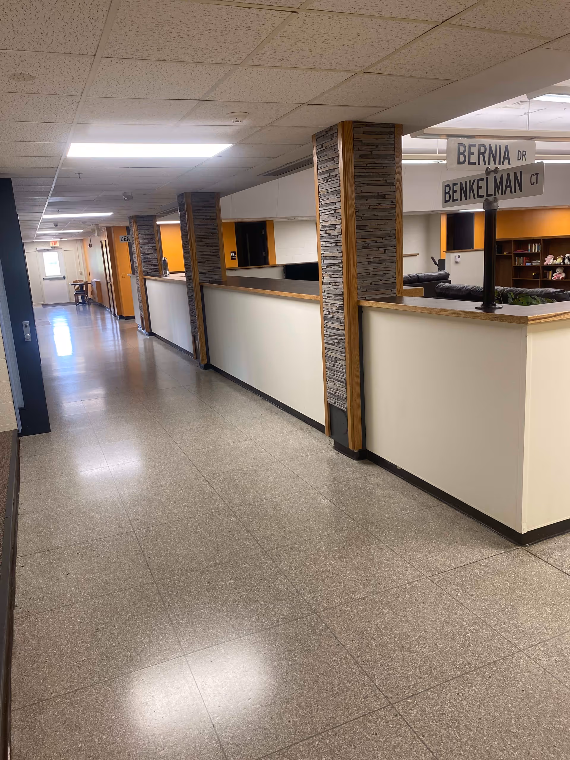 A long indoor hallway with tiled flooring and a drop ceiling with fluorescent lights. On the right side, there are half walls with stone and wood pillars, and a street sign post with signs reading 'BERNIA DR' and 'BENKELMAN CT'. In the background, there is a seating area with couches and shelves.