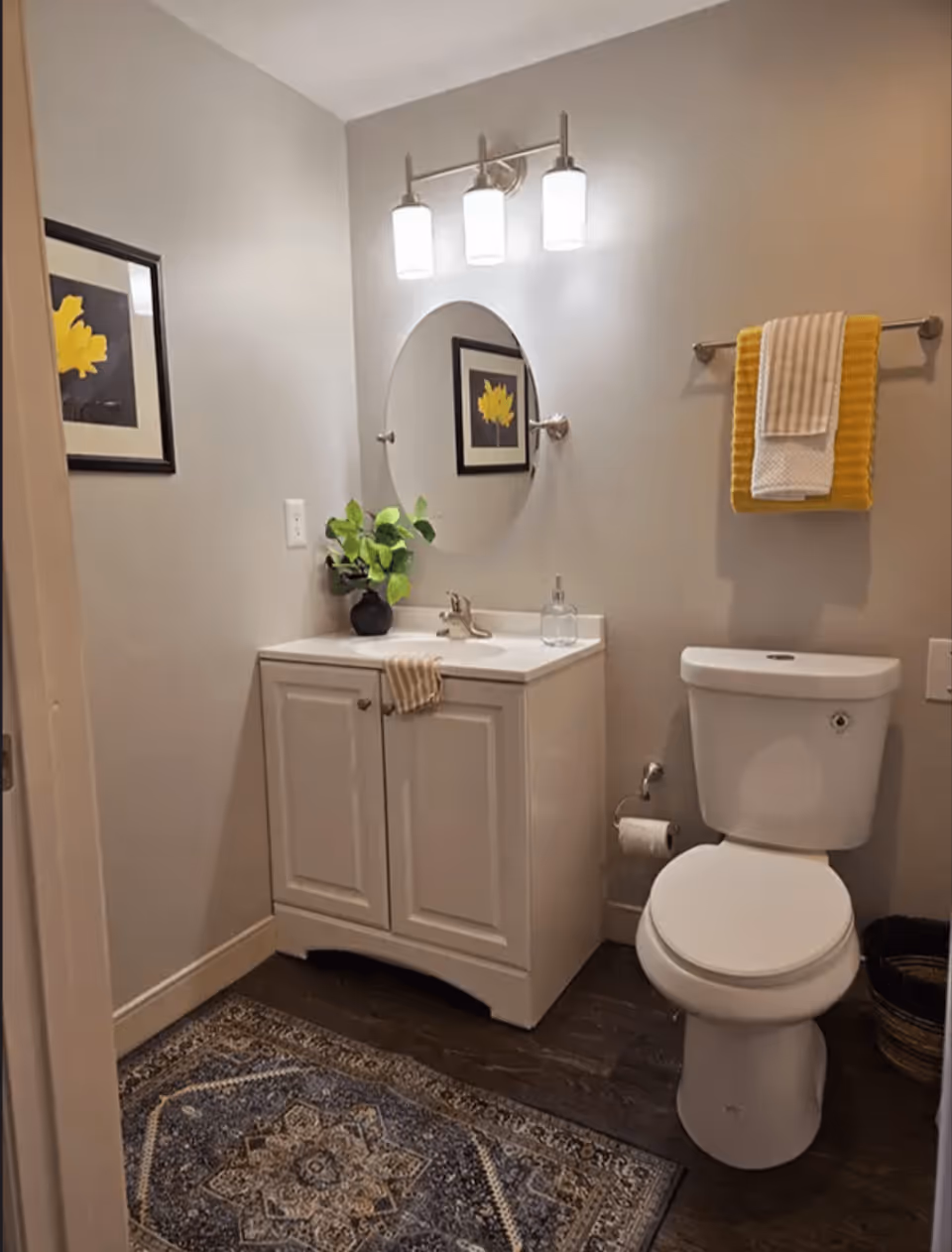 Bathroom interior showing a white vanity with a round mirror, toilet, towel rack with yellow towels, and a patterned floor rug.