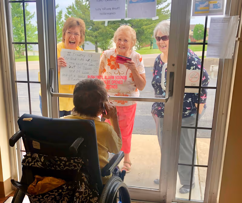 A person in a wheelchair inside a building is talking on the phone while three elderly women stand outside the glass door smiling and holding a handwritten sign. The door has a red warning text about the alarm sounding if pushed. Trees and a parking lot are visible outside.