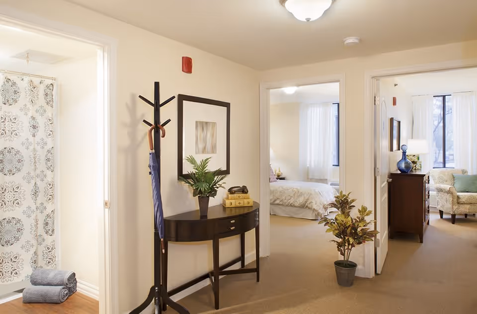 Interior view of a senior living facility showing a hallway with a coat rack holding an umbrella, a small table with a plant and books, and two open doorways. One doorway leads to a bedroom with a bed and window, and the other doorway leads to a living room area with a chair, lamp, and window. A bathroom with a patterned shower curtain and folded towels is visible on the left.