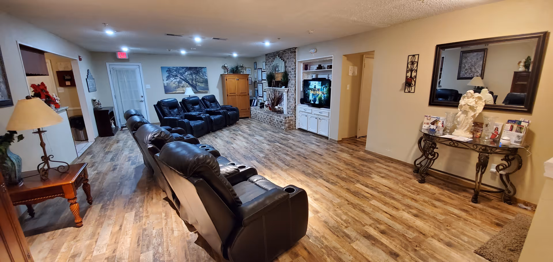 A spacious living room with wooden flooring, featuring two rows of black leather recliner chairs facing a brick fireplace and a TV mounted in a white built-in cabinet. The room has beige walls, a large mirror above a decorative table with various items, a floor lamp on a wooden side table, and framed artwork on the walls.