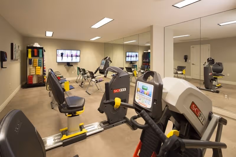 A fitness room with exercise equipment including SciFit machines, chairs, and a rack of colorful exercise mats and weights. A large mirror covers one wall, and a television screen is mounted on the opposite wall displaying a workout video.