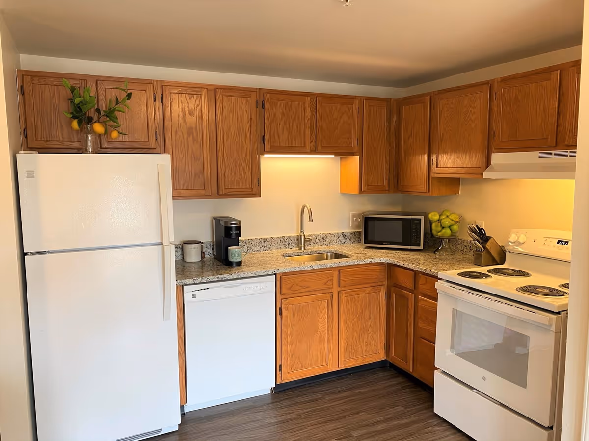 Compact kitchen with oak cabinets, white refrigerator, dishwasher and stove, granite countertops, sink, microwave, and fruit accents.