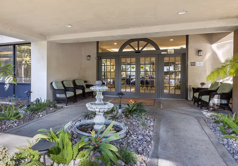 Covered entrance area of a senior living facility with a stone fountain surrounded by plants and rocks in the center. There are wicker chairs with green cushions on both sides near the glass double doors leading inside the building.