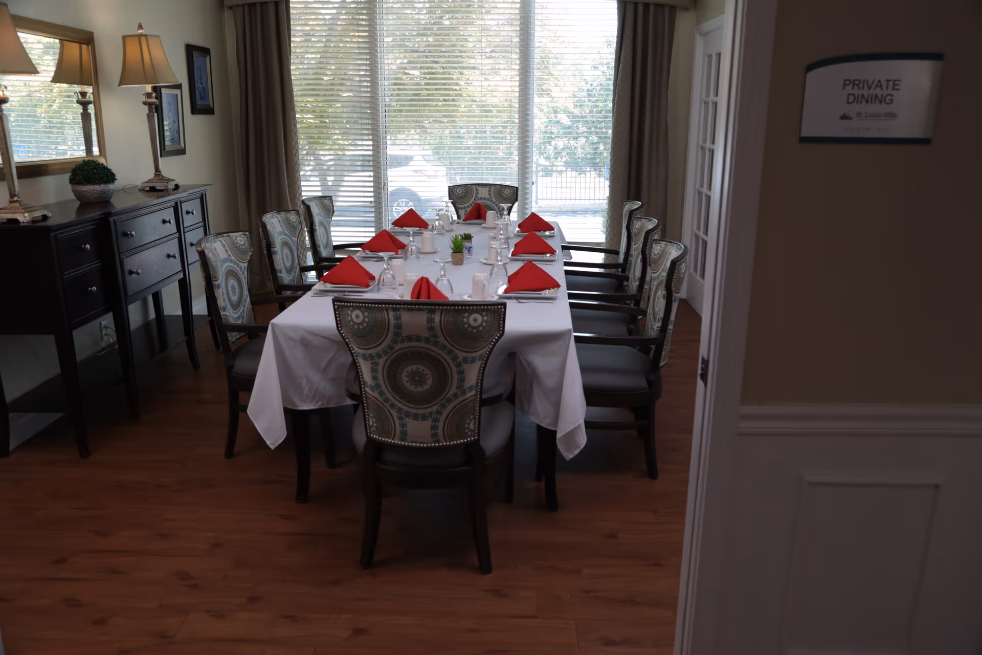 A private dining room with a rectangular table covered with a white tablecloth, set with red folded napkins, glasses, and white cups. The room has eight upholstered chairs with patterned backs, wooden flooring, a sideboard with two lamps and a small plant, and large windows with blinds letting in natural light.
