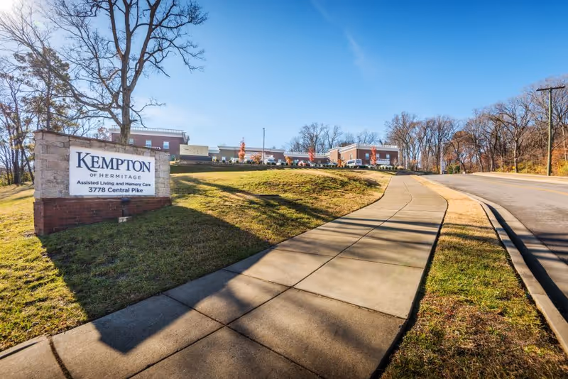 Front exterior of Kempton of Hermitage assisted living with a sign by a sidewalk leading up to the building