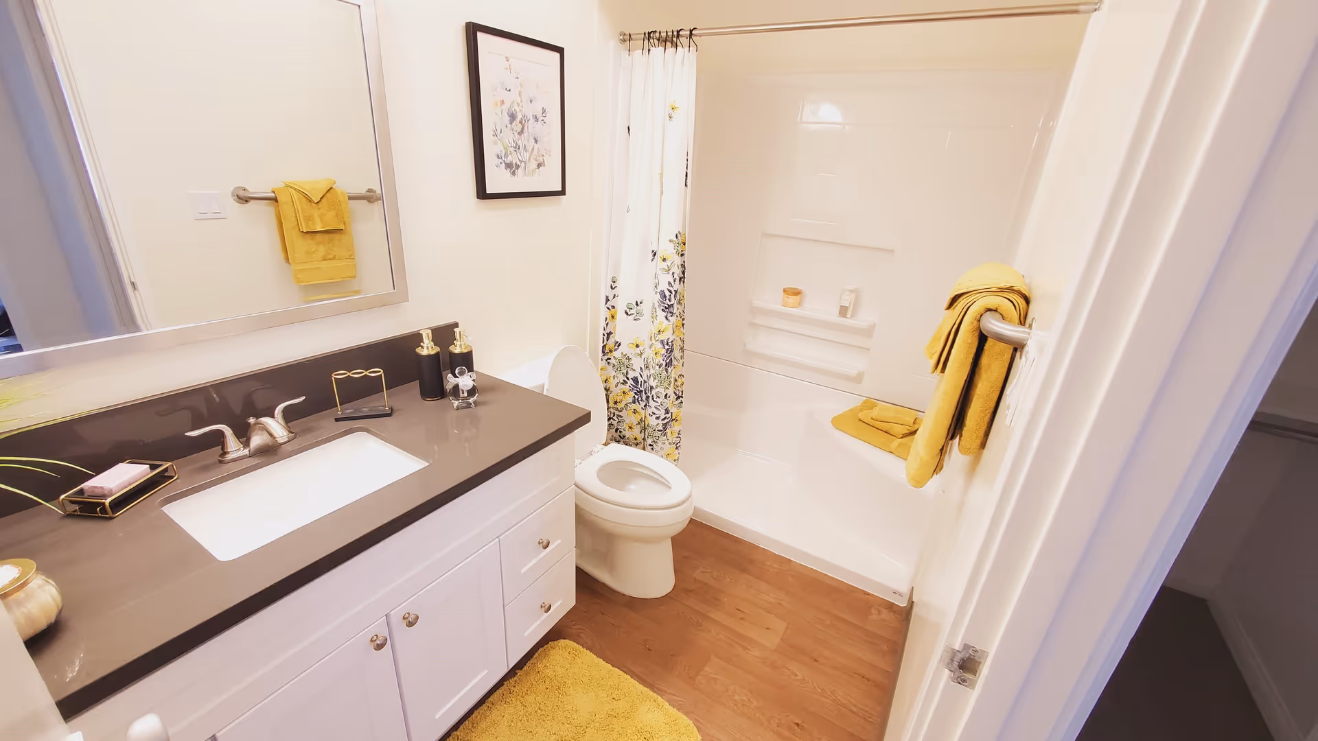 A clean bathroom featuring a white vanity with a dark countertop and a built-in sink, a large mirror above the sink, a toilet, and a bathtub with a floral shower curtain. Yellow towels and a matching bath mat add color to the space, which has light-colored walls and wood flooring.