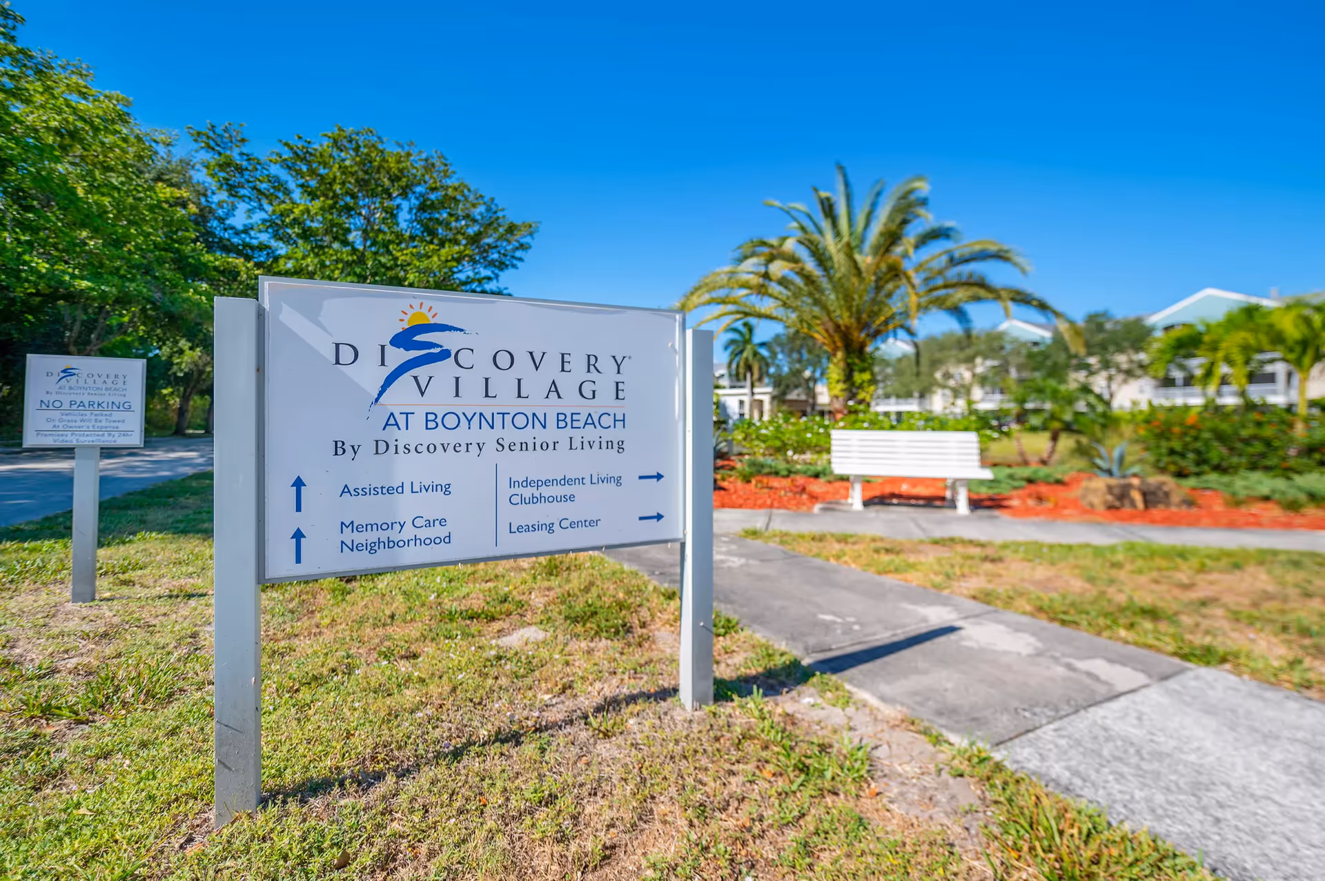 Entrance sign for Discovery Village at Boynton Beach on landscaped grounds with a white bench, palm trees, and buildings in the background.