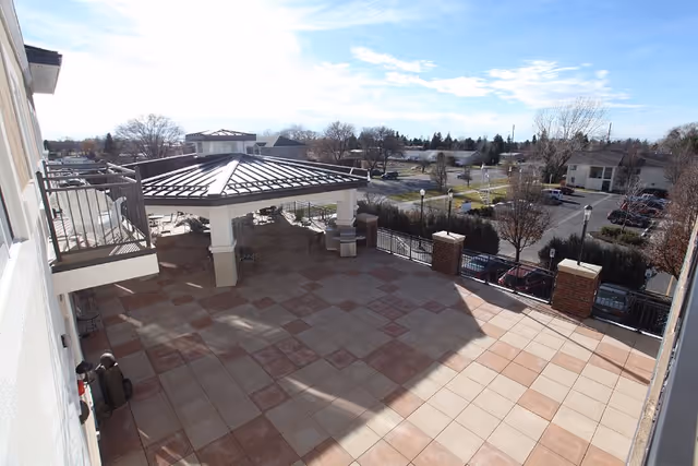 Outdoor patio area with a tiled floor and a covered pavilion with a metal roof. The patio has a railing along the edge and overlooks a parking lot and trees in the background under a partly cloudy sky.