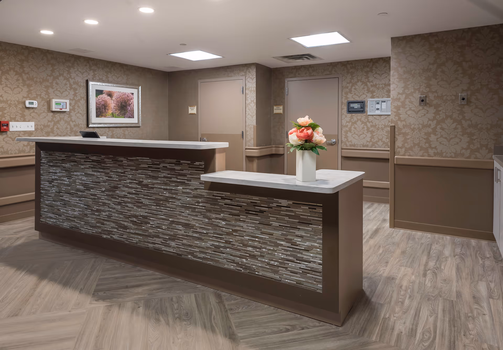 Reception desk in a senior living facility lobby with a vase of flowers, patterned wallpaper and two closed doors.