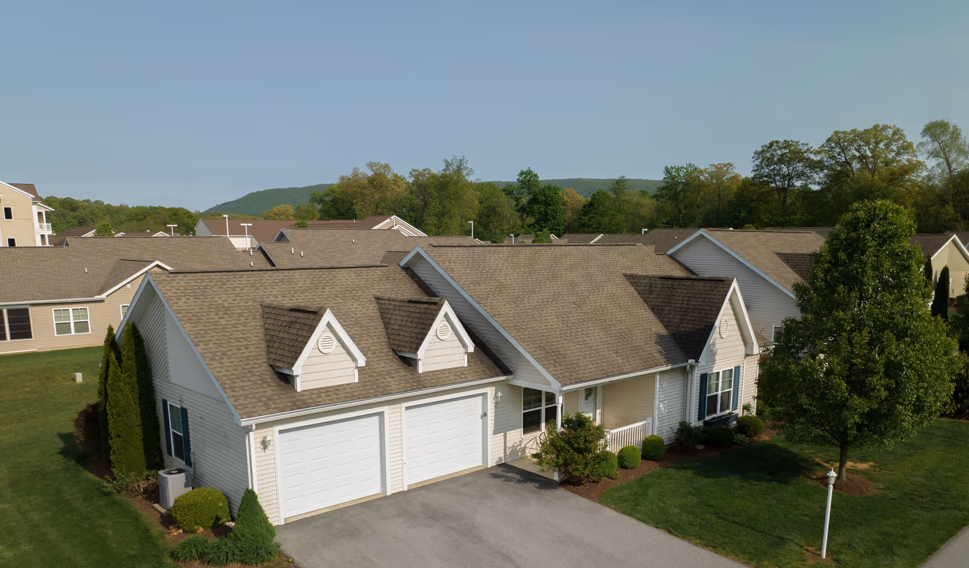Aerial view of connected single-story residential units with an attached double garage, front porch, and landscaped lawn with trees.