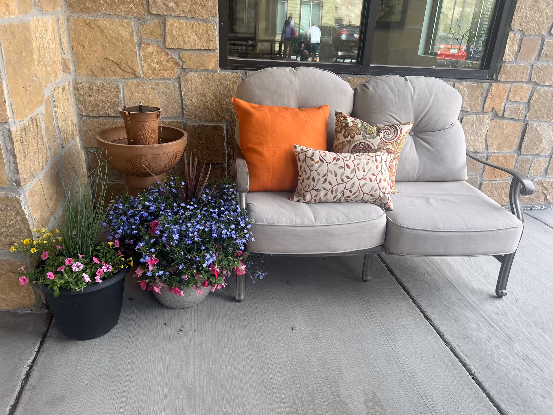 Outdoor seating area with a cushioned metal loveseat featuring three decorative pillows in orange and patterned designs. Next to the loveseat are two flower pots with colorful flowers and a small decorative water fountain. The background shows a stone wall and a window reflecting people and cars outside.