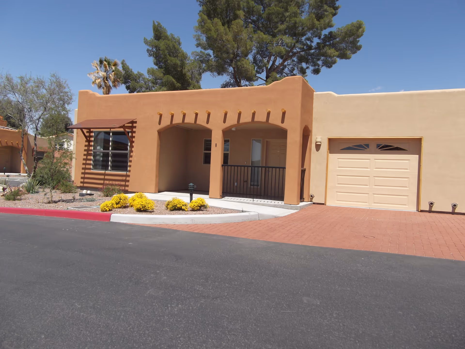 Exterior view of a single-story southwestern style building with tan stucco walls, a covered porch with arches, a garage with a beige door, desert landscaping with yellow flowering bushes, and a clear blue sky.