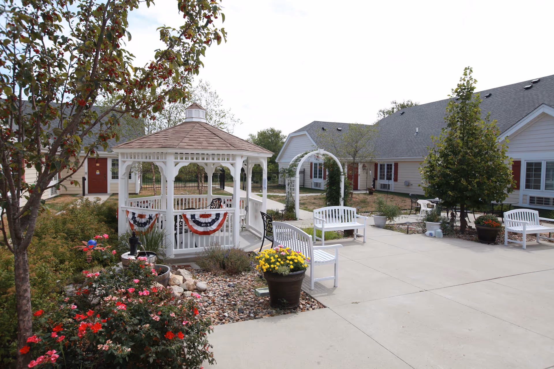 Outdoor courtyard area of a senior living facility featuring a white gazebo decorated with red, white, and blue bunting. There are white benches, potted flowers, and a small garden with rocks and blooming plants. The background shows single-story buildings with gray roofs and red doors and shutters.