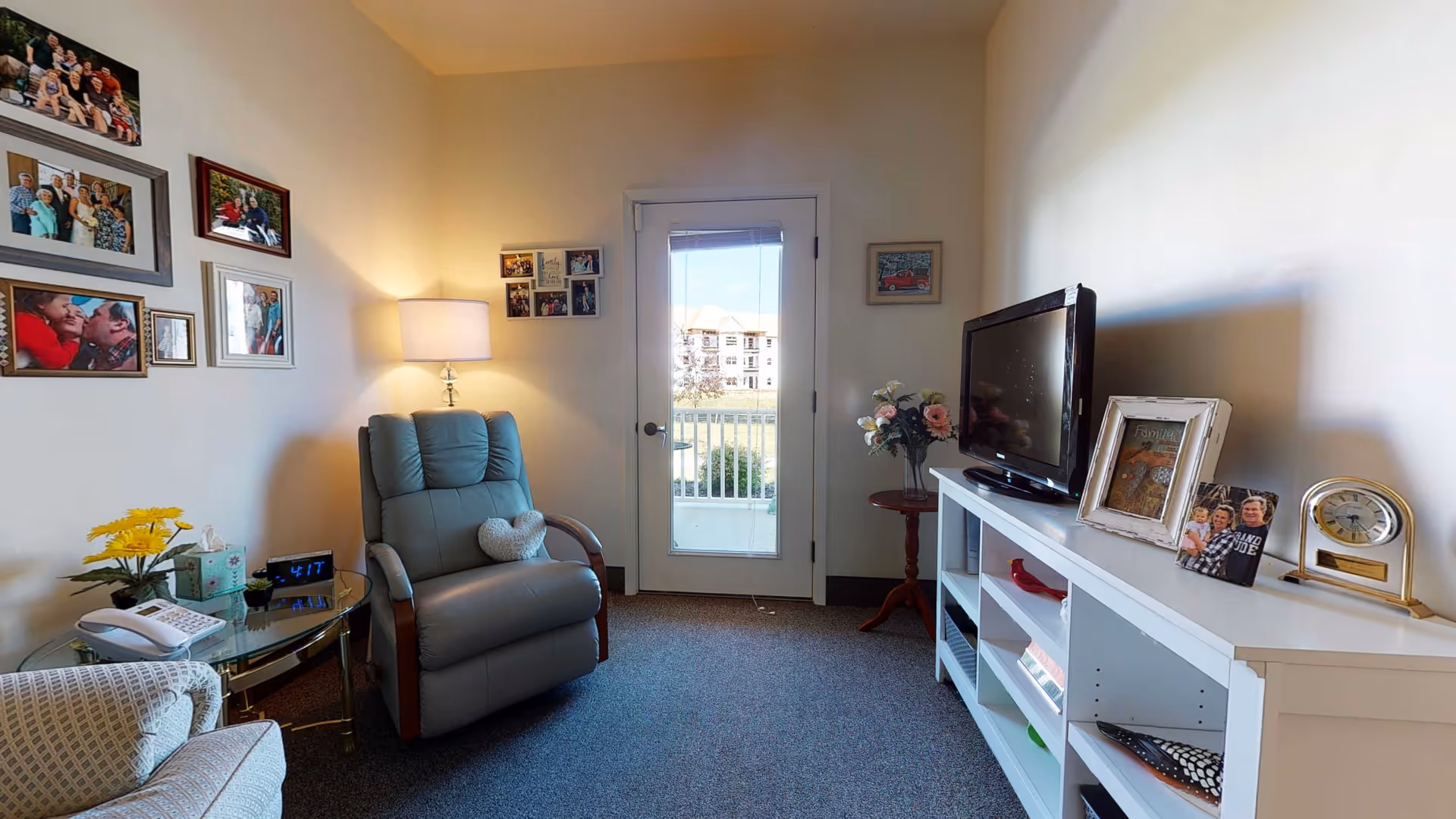 Small living room with a recliner, TV on a white console, framed family photos, and a glass door leading to a balcony.