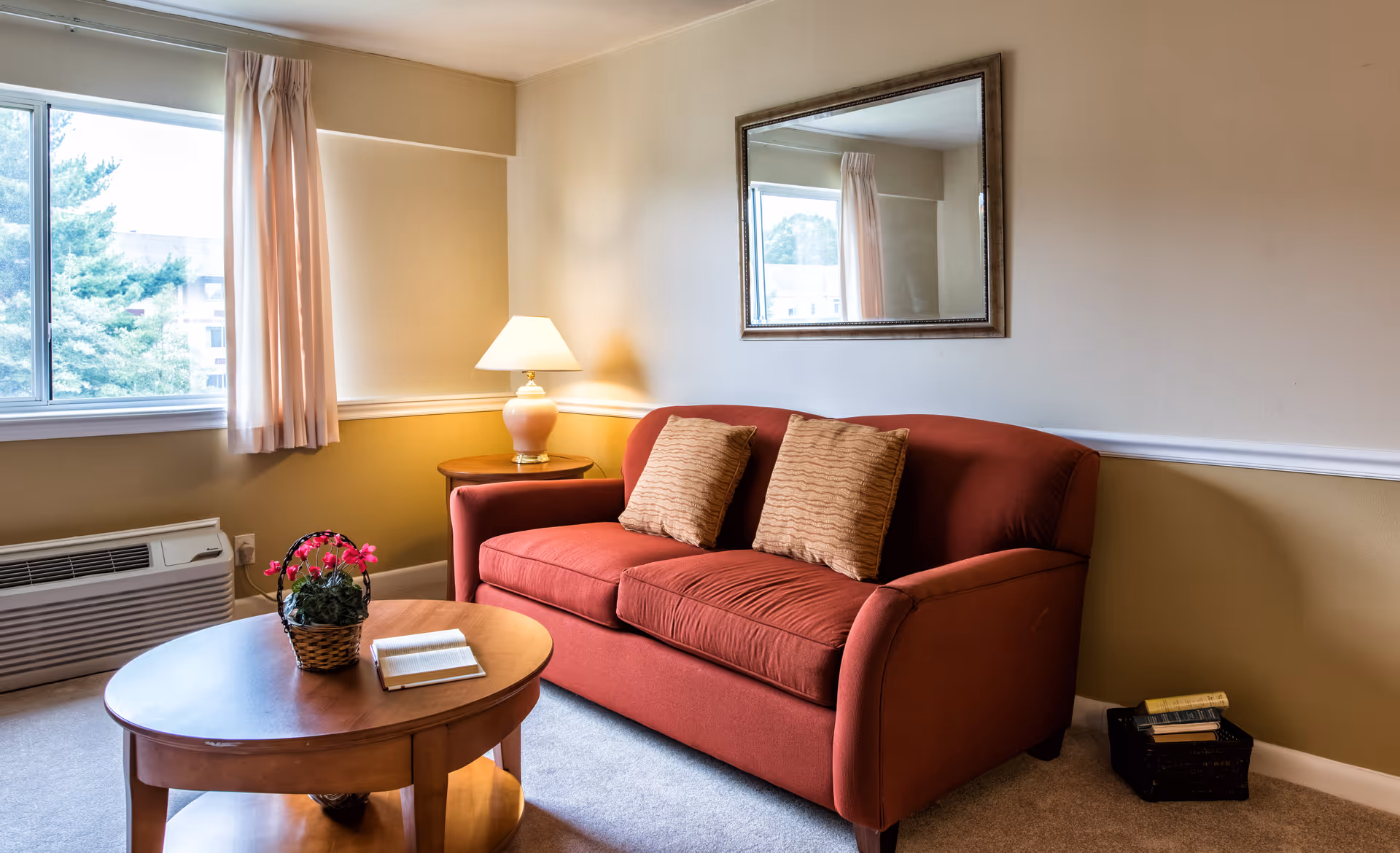 A cozy living room area with a red sofa adorned with two beige cushions, a wooden coffee table with a basket of pink flowers and an open book, a side table with a lamp, a large window with beige curtains, and a large mirror on the wall reflecting part of the room.