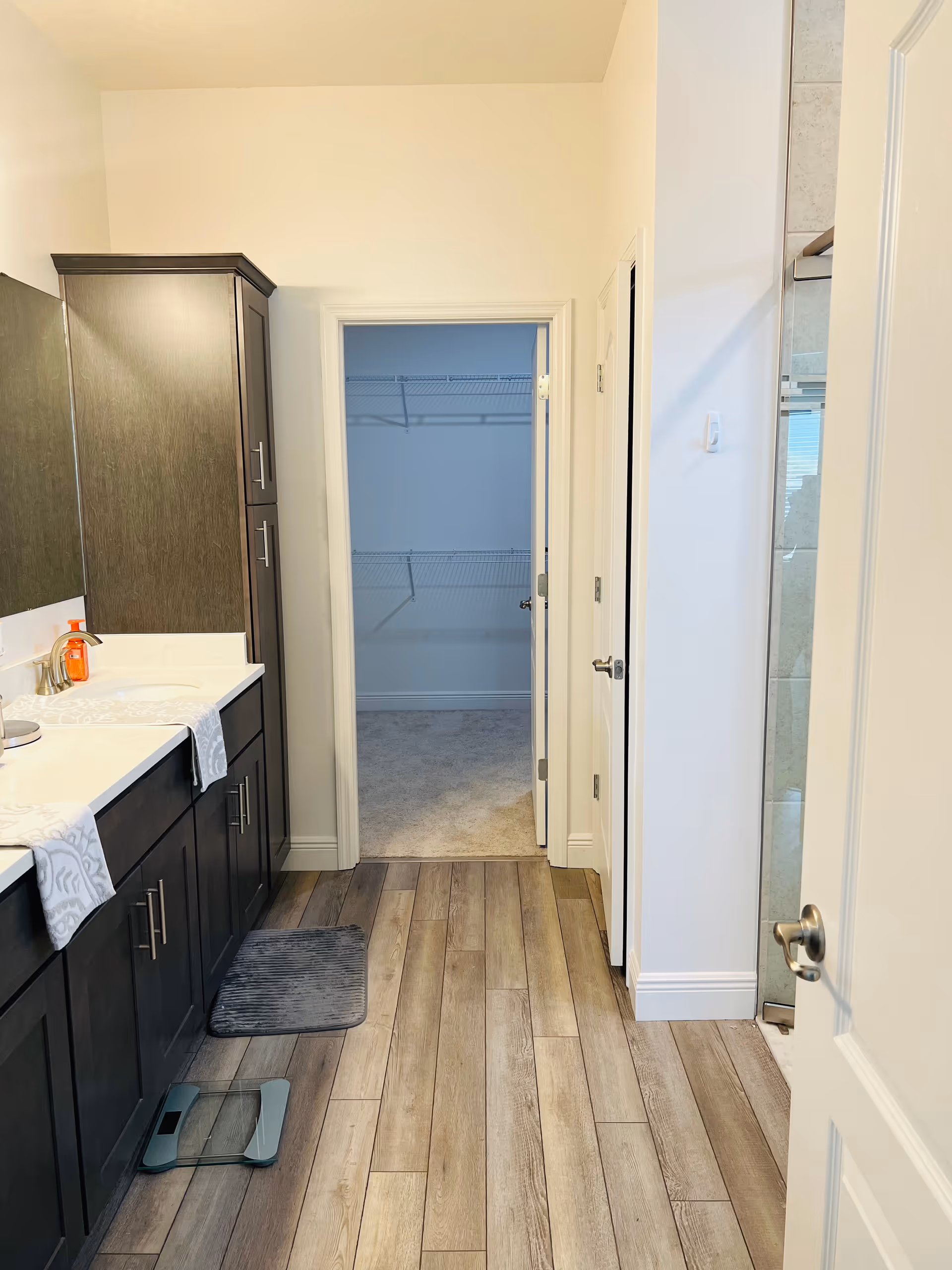 Interior view of a bathroom with wooden floor tiles, dark wood cabinets with a white countertop, a sink with a soap dispenser, a gray bath mat, and a glass shower door on the right. There is an open doorway leading to a walk-in closet with wire shelving.